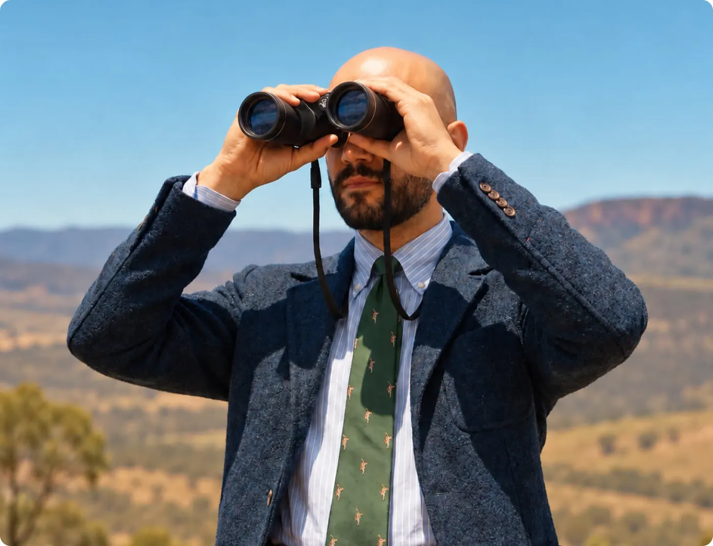 Bald, bearded man in a blazer and tie looking through binoculars, hills and scrub behind him