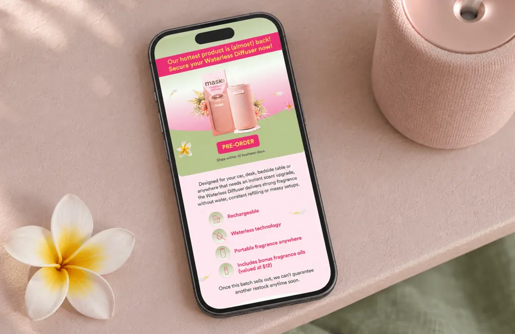 Phone on a pink table showing a MaskCo email to pre-order the Waterless Diffuser, beside a frangipani
