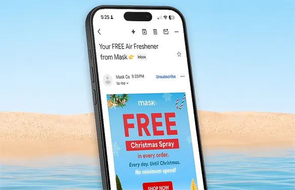 Phone inbox open on a MaskCo email for a free Christmas spray, in front of a beach and calm sea