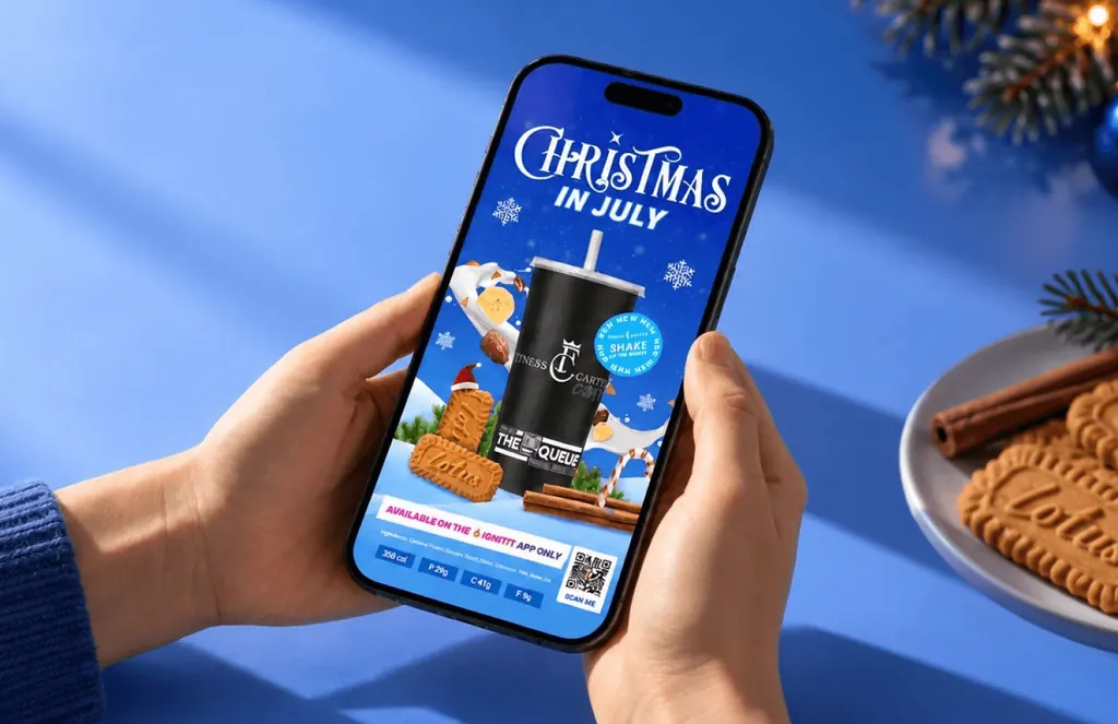 Hand holding a phone with a Fitness Cartel Christmas in July shake promo, beside a plate of Lotus biscuits