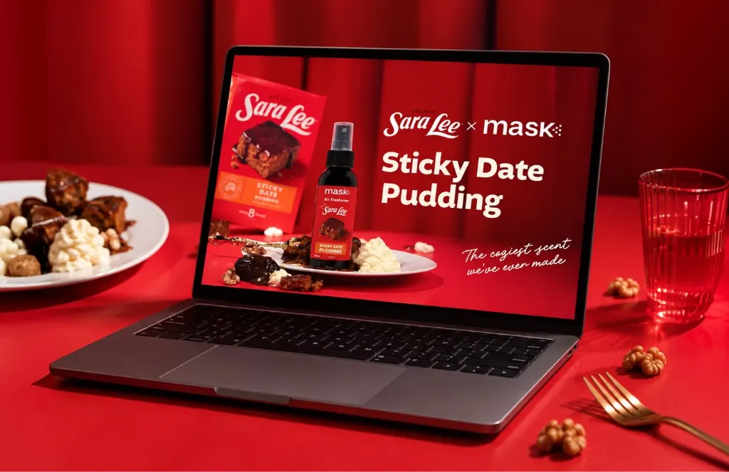 Laptop on a red table showing the Sara Lee x MaskCo air freshener ad, beside a plate of pudding and cream