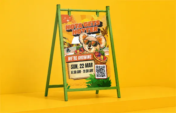 Green A-frame sign holding the HOTA Gets Hotter koala poster with date and QR code, against a yellow wall