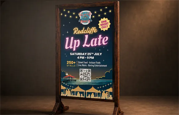 Wooden A-frame with a neon-style Redcliffe Up Late poster of market stalls by the water at night